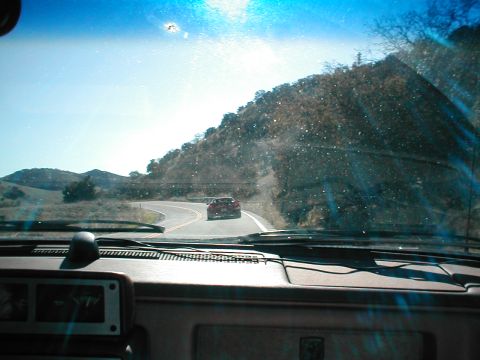 1986 Pontiac Fiero - View from inside a vehicle looking at a red sports car ahead on a scenic, winding road.