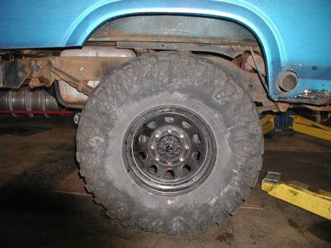 1985 Ford Bronco - A close-up side view of the rear wheel and suspension of a blue Ford Bronco. The vehicle has large, aggressive off-road tires and appears t…