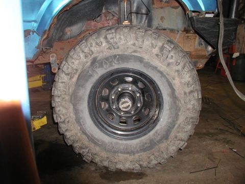 1985 Ford Bronco - A close-up view of a large, knobby off-road tire mounted on a black rim. The tire is on a vehicle with a blue fender and significant rust v…