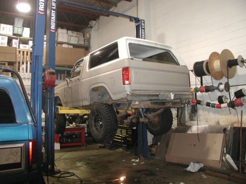 1985 Ford Bronco - A light-colored Ford Bronco is raised on a hydraulic lift in a garage. The vehicle is viewed from the rear quarter angle, showing its lifte…