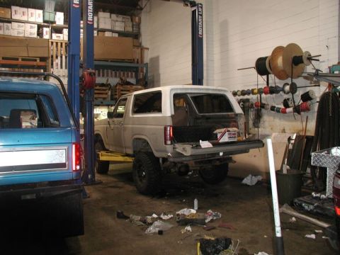 1985 Ford Bronco - A white and tan Ford Bronco is lifted on a car lift in a garage. The rear hatch is open, revealing the cargo area and some tools.