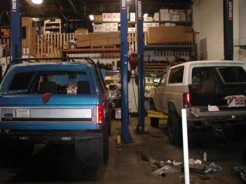 1985 Ford Bronco - Two Ford Broncos are parked in a garage with car lifts. The blue Bronco is in the foreground, viewed from the rear, and a tan Bronco is to…