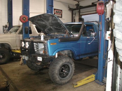 1985 Ford Bronco - A blue Ford Bronco with its hood open is parked on a lift in a garage, next to a lighter colored pickup truck. The Bronco has a snorkel, a…
