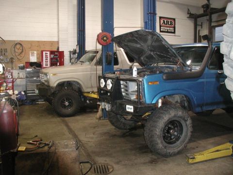 1985 Ford Bronco - Two modified Ford Broncos are parked in a garage, with the blue Bronco in the foreground having its hood open and snorkel installed. The ta…