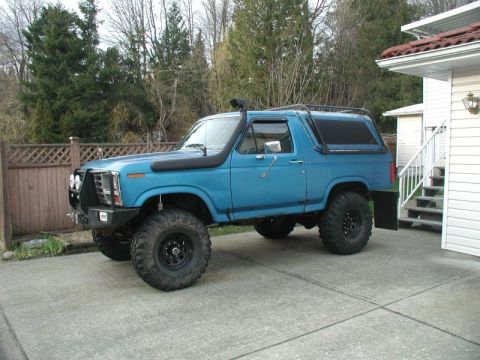 1985 Ford Bronco - A lifted, bright blue 1980s Ford Bronco with a snorkel and roof rack is parked on a concrete driveway next to a house.