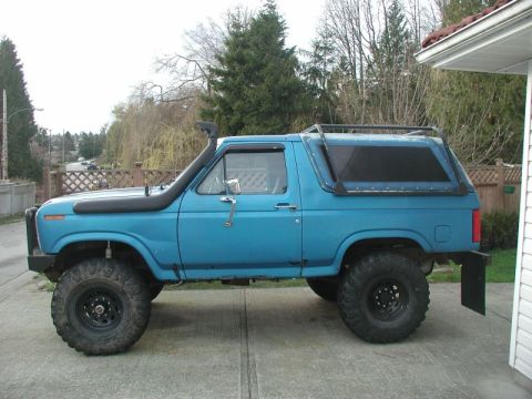 1985 Ford Bronco - Side profile of a lifted, blue 1980s Ford Bronco with a snorkel, roof rack, and large off-road tires parked on a concrete driveway.
