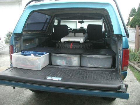 1985 Ford Bronco - Cargo area of a teal-blue Ford Bronco photographed with the tailgate down, showing a raised flat load floor built over three clear plastic…