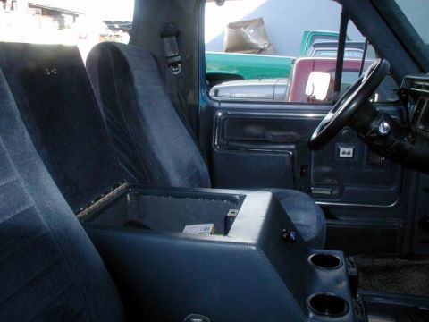 1985 Ford Bronco - Truck interior shot from the rear seat showing a custom blue center console with its lid propped open, carpeted storage bin, twin molded cu…
