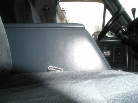 1985 Ford Bronco - Low-angle interior shot along a blue cloth bench seat showing the smooth blue vinyl side of a center console with its chrome pull latch. Th…