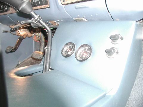 1985 Ford Bronco - Driver-side face of a custom blue vinyl center console fitted with two chrome-bezel aftermarket gauges and two toggle switches. The bent sh…