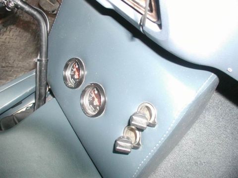 1985 Ford Bronco - Detail of two small black-face aftermarket gauges with red needles and two chrome rocker switches mounted in the blue vinyl side panel of a…