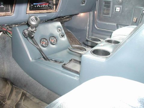 1985 Ford Bronco - Interior view from the passenger side of a blue vinyl-wrapped center console with two round Auto Meter gauges, two chrome pull switches, an…