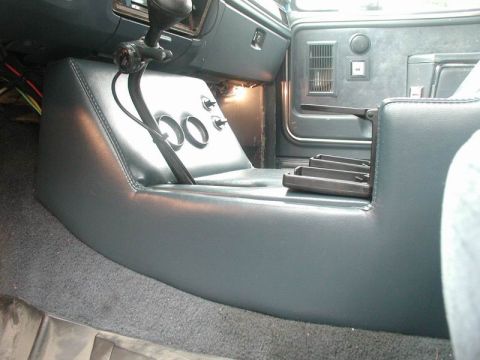 1985 Ford Bronco - Passenger-side view of a blue vinyl-upholstered center console in progress, with two empty round gauge holes cut into the angled face and t…