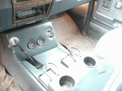 1985 Ford Bronco - Downward view of a finished blue vinyl center console with two Auto Meter gauges, two chrome pull switches, an automatic shifter with patte…