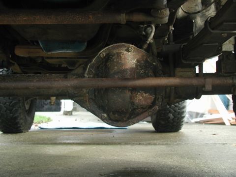 1985 Ford Bronco - The underside of a vehicle shows a rusty differential housing and leaf springs. The vehicle has large off-road tires visible on either side.