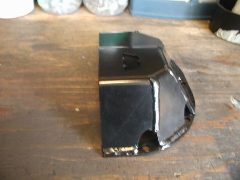 1985 Ford Bronco - A black metal part, possibly a bracket or shield, is shown on a wooden surface. The part has visible welds and mounting holes.