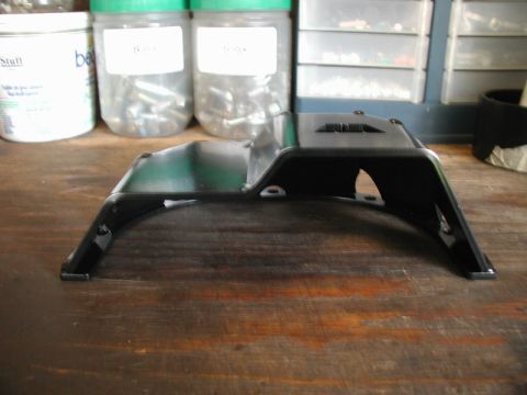 1985 Ford Bronco - A black metal part, possibly a skid plate or bracket, is shown from a side angle resting on a wooden surface. In the background, several cl…