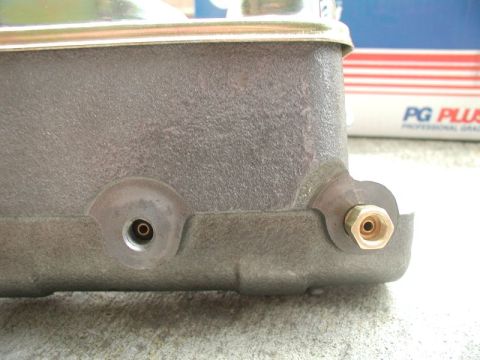 1985 Ford Bronco - Close-up side view of a gray metal brake master cylinder with two brass fittings. A portion of a box with red and white stripes and the tex…