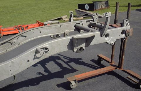 1985 Ford Bronco - A bare, unpainted vehicle frame, possibly for a truck or SUV, is supported by metal stands and dollies on an asphalt surface with grass in…
