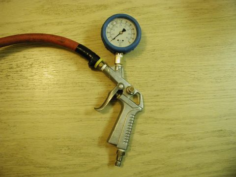 1985 Ford Bronco - A close-up view of an air compressor blow gun with a pressure gauge attached, connected to a red hose. The tool is resting on a light-color…