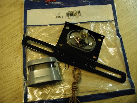 1985 Ford Bronco - A Grote license plate light assembly kit is shown laid out on a wooden surface. The kit includes a black metal bracket with a bulb, a gray…