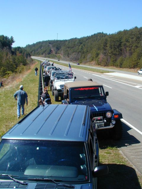 1988 Ford Bronco - A convoy of Jeeps is stopped on the side of a highway, with a blue Jeep in the foreground and a line of others behind it.