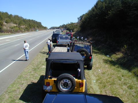 1988 Ford Bronco - A line of Jeeps and other vehicles are parked on the shoulder of a highway on a sunny day.