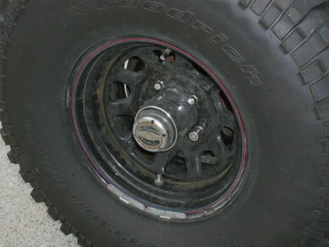 1985 Ford Bronco - A close-up view of a black BFGoodrich All-Terrain T/A tire mounted on a black steel wheel with a red and blue stripe accent. The wheel feat…
