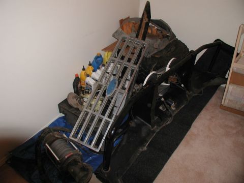 1985 Ford Bronco - A collection of vehicle parts is stored in a corner of a room, including a large chrome grille with a blue emblem, a winch, and what appear…