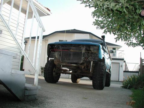 1985 Ford Bronco - The rear of a lifted, blue 1985 Ford Bronco is parked in a driveway next to a white house. The vehicle has large off-road tires and a snork…