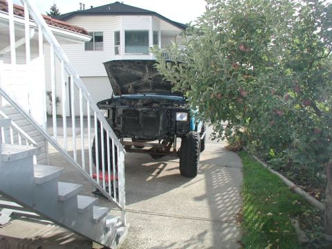 1985 Ford Bronco - A blue 1985 Ford Bronco with its hood open is parked on a concrete driveway next to an apple tree. An outdoor staircase is visible in the f…