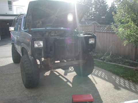 1985 Ford Bronco - A dark blue Ford Bronco with the hood open and the front grille removed is parked in a driveway. The vehicle has large off-road tires and a…