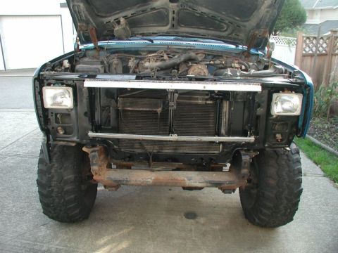 1985 Ford Bronco - The front view of a teal Ford Bronco with its hood open, revealing the engine bay. The vehicle's grille, headlights, and large off-road tir…