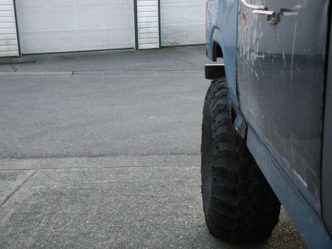 1985 Ford Bronco - A close-up, side view of the rear fender and large off-road tire of a blue vehicle. The vehicle appears to be parked on asphalt next to a c…