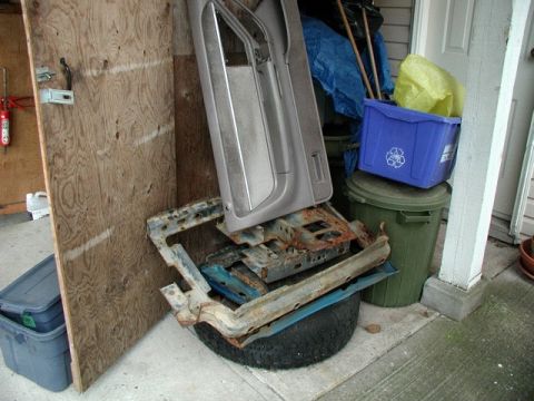 1985 Ford Bronco - A collection of vehicle parts, including a car door, a car seat frame, and a tire, are stored outdoors next to a plywood partition. A green…