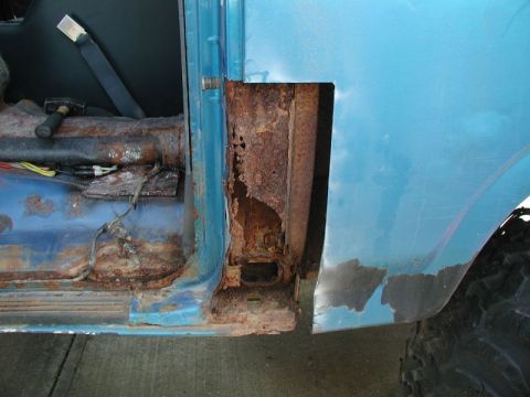 1985 Ford Bronco - The interior driver's side floor and door jamb of a blue vehicle show extensive rust damage. A hammer rests on the rusted floor near expose…