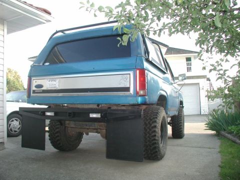1985 Ford Bronco - The rear of a lifted, blue Ford Bronco with large mud tires and black mud flaps is parked in a driveway. The vehicle has a custom rear bump…