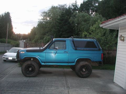 1985 Ford Bronco - A lifted, teal 1980s Ford Bronco with a snorkel and roof rack is parked on a concrete driveway next to a white house. A white sedan is park…