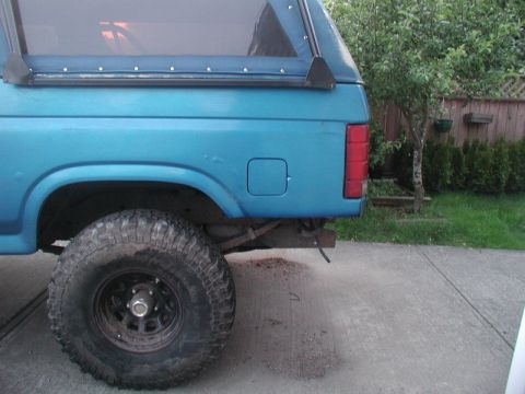 1985 Ford Bronco - The rear quarter panel and large off-road tire of a blue SUV are visible. The vehicle has a soft top with visible snaps along the top edge.