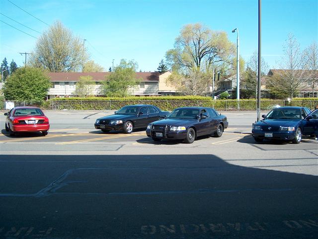Four sedans parked in a lot, including a red sedan, a black sedan, a dark blue sedan, and a lighter blue sedan. The dark blue sedan is a 2001 Ford Crown Victoria
