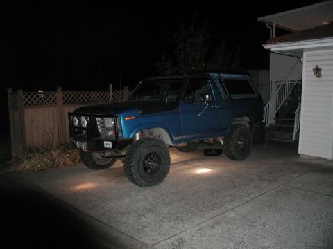 1985 Ford Bronco - A lifted blue 1980s Ford Bronco is parked on a concrete driveway at night. The vehicle has large off-road tires and a brush guard on the fr…