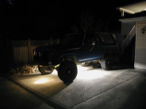 1985 Ford Bronco - A dark blue Ford Bronco is parked on a concrete driveway at night, illuminated by bright headlights. The vehicle has large off-road tires a…
