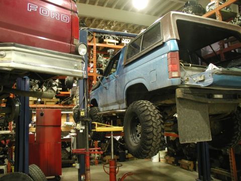 1985 Ford Bronco - A blue Ford Bronco is lifted on a car lift in a garage with shelves of vehicle parts in the background. A red Ford pickup truck is also par…