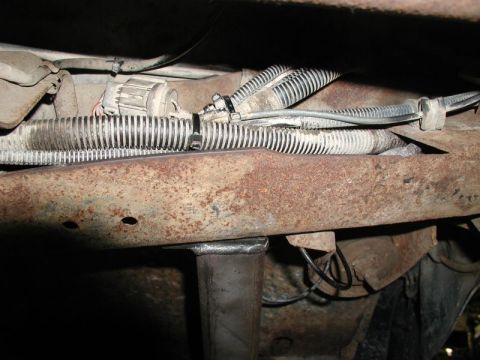 1985 Ford Bronco - The underside of a vehicle shows rusty metal framing with various electrical wires and conduits secured with zip ties. A metal support brac…