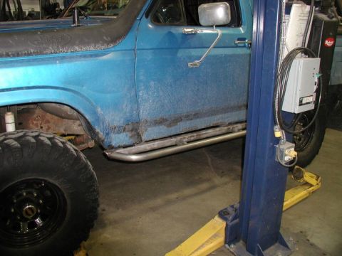 1985 Ford Bronco - The side view of a blue Ford Bronco is shown, lifted on a blue and yellow car lift. Visible modifications include a snorkel and custom meta…