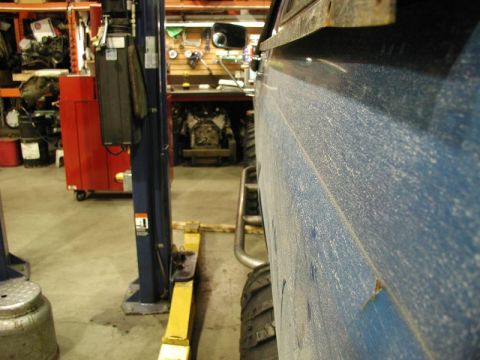 1985 Ford Bronco - Close-up side view of a dusty blue vehicle, possibly a 1985 Ford Bronco, parked in a garage with a vehicle lift and engine parts visible in…