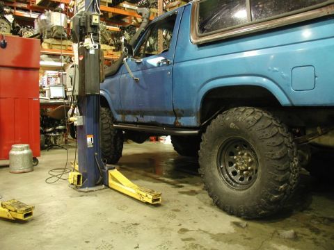 1985 Ford Bronco - A blue Ford Bronco is parked on a lift in a garage with shelves of car parts in the background. The vehicle has large off-road tires, a sno…