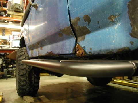 1985 Ford Bronco - A close-up side view of a rusty blue Ford Bronco's lower body panel and a black tubular side step. The vehicle is in a workshop setting wit…
