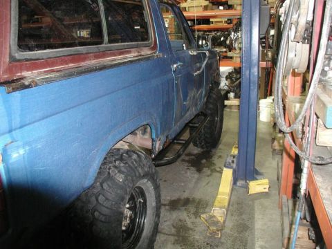 1985 Ford Bronco - A blue Ford Bronco with large off-road tires and custom rock sliders is parked on a lift in a garage. The vehicle shows signs of wear and t…