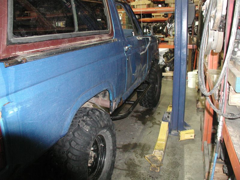 Blue 1985 Ford Bronco with off-road tires and rock sliders on a lift.